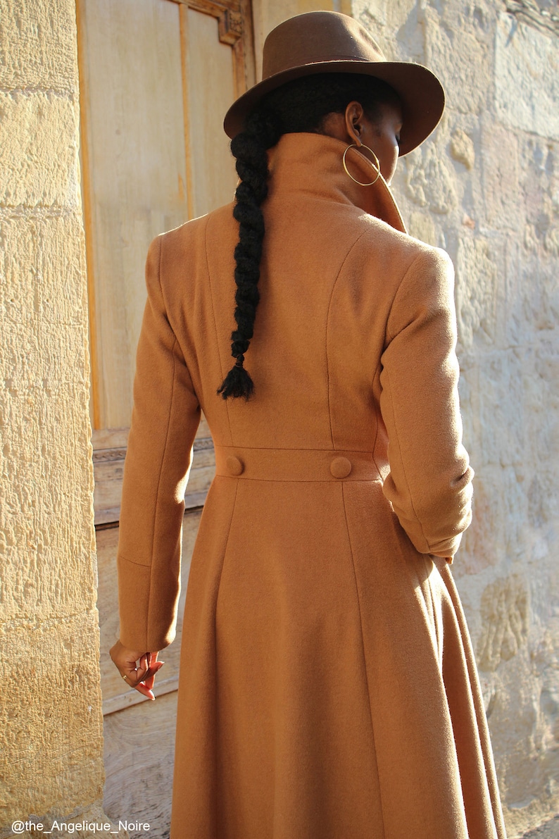 May include: A brown long coat with a high collar and a fitted waist. The coat has a long skirt and two buttons on the back. The person is wearing a brown hat.