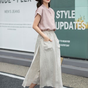 May include: A woman wearing a pink short-sleeved shirt, a beige linen skirt with a side slit, and brown wedge sandals. She is also wearing a brown hat with a black ribbon and a pair of earrings. The background is a wall with the text "Introducing Aude - Flex Men's Jeans" and "Style Updates".