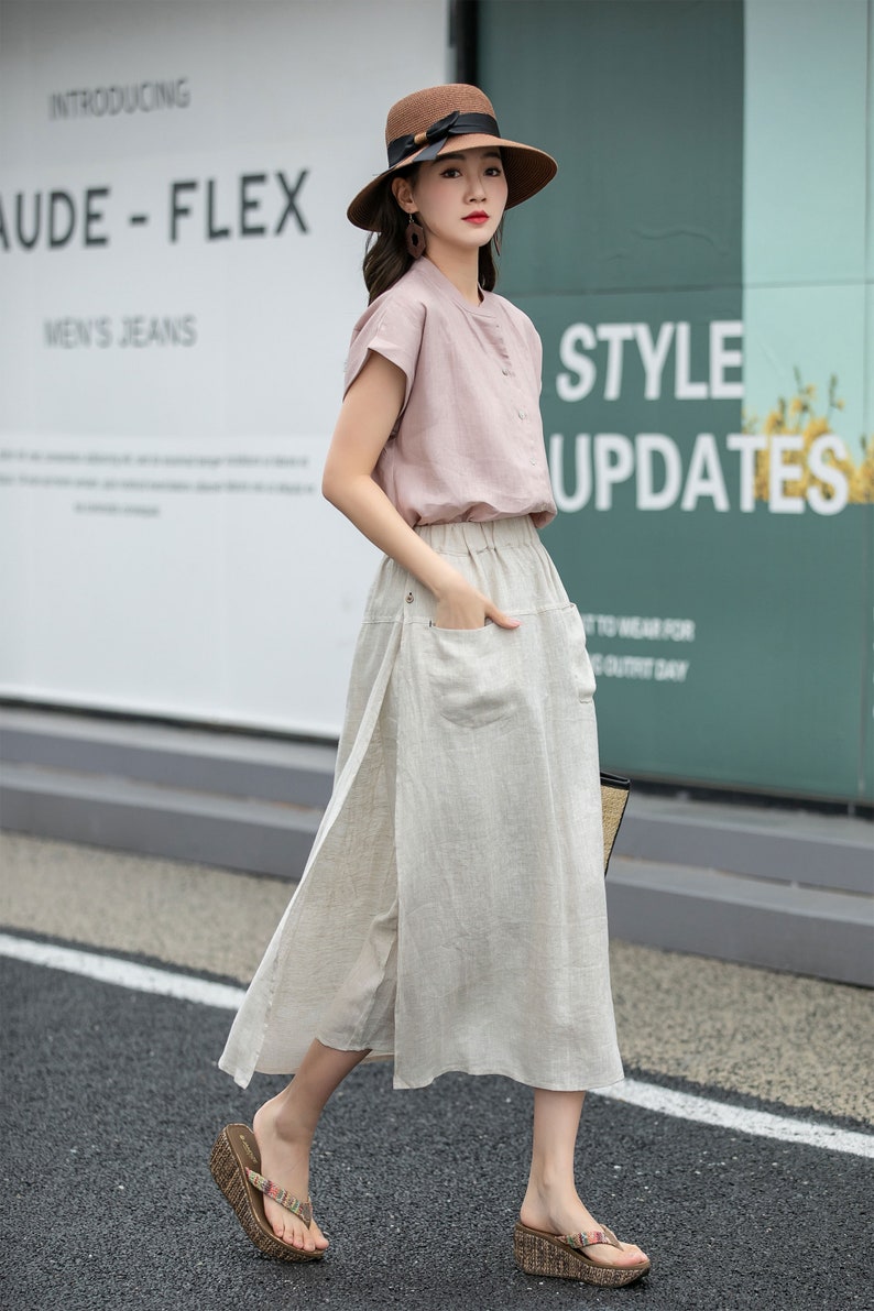 May include: A woman wearing a pink short-sleeved shirt, a beige linen skirt with a side slit, and brown wedge sandals. She is also wearing a brown hat with a black ribbon and a pair of earrings. The background is a wall with the text "Introducing Aude - Flex Men's Jeans" and "Style Updates".