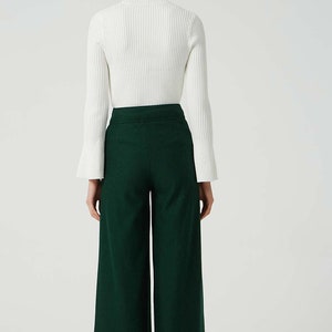 May include: A woman wearing a white turtleneck sweater and dark green wide-leg trousers. The trousers have a high waist and a wide leg.