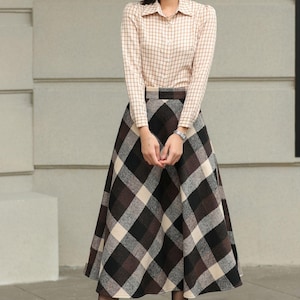 May include: A woman wearing a beige plaid button-down shirt and a long plaid skirt in shades of brown, black, and cream. She is also wearing brown lace-up boots. The outfit is a classic, vintage-inspired style.