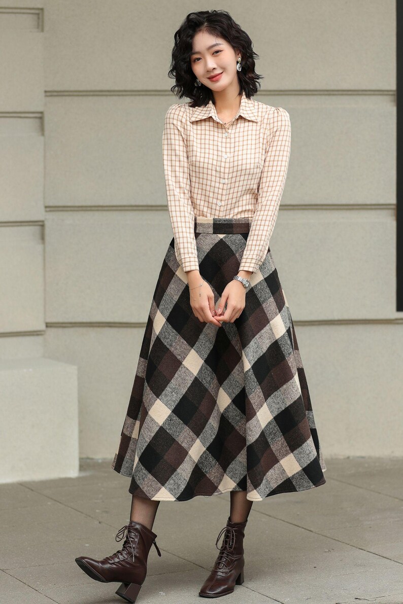 May include: A woman wearing a beige plaid button-down shirt and a long plaid skirt in shades of brown, black, and cream. She is also wearing brown lace-up boots. The outfit is a classic, vintage-inspired style.