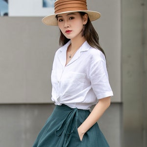 May include: A woman wearing a white linen shirt with a knotted front and a dark green wrap skirt. She is wearing a wide-brimmed straw hat with a brown band.
