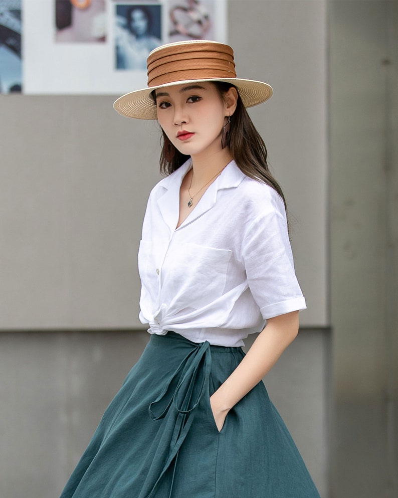 May include: A woman wearing a white linen shirt with a knotted front and a dark green wrap skirt. She is wearing a wide-brimmed straw hat with a brown band.