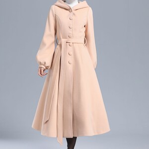 May include: A long, light pink hooded coat with a belted waist and button closure. The coat has a flared skirt and long sleeves.