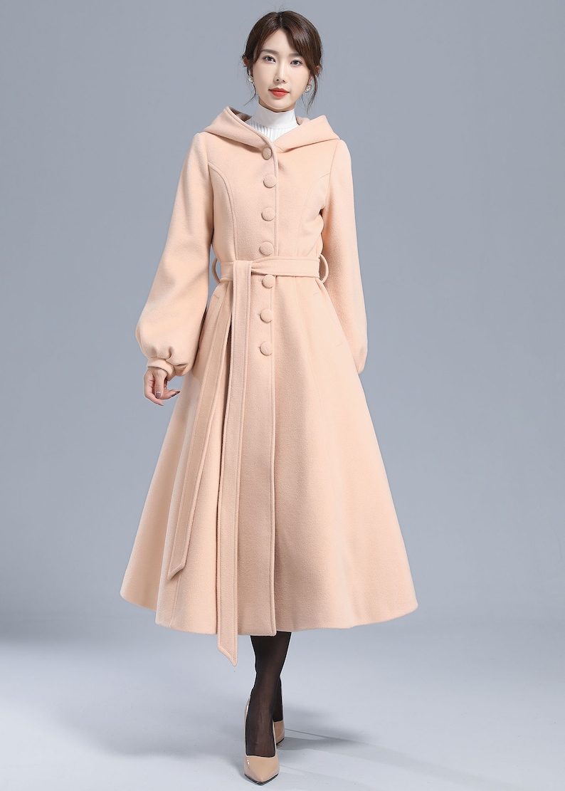 May include: A long, light pink hooded coat with a belted waist and button closure. The coat has a flared skirt and long sleeves.