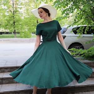 Puede incluir: Un vestido verde oscuro con escote barco y mangas cortas, que presenta un corpi&ntilde;o ajustado y una falda amplia y acampanada. El conjunto incluye un sombrero de sol blanco de ala ancha, guantes beige largos y zapatos de sal&oacute;n blancos cerrados.