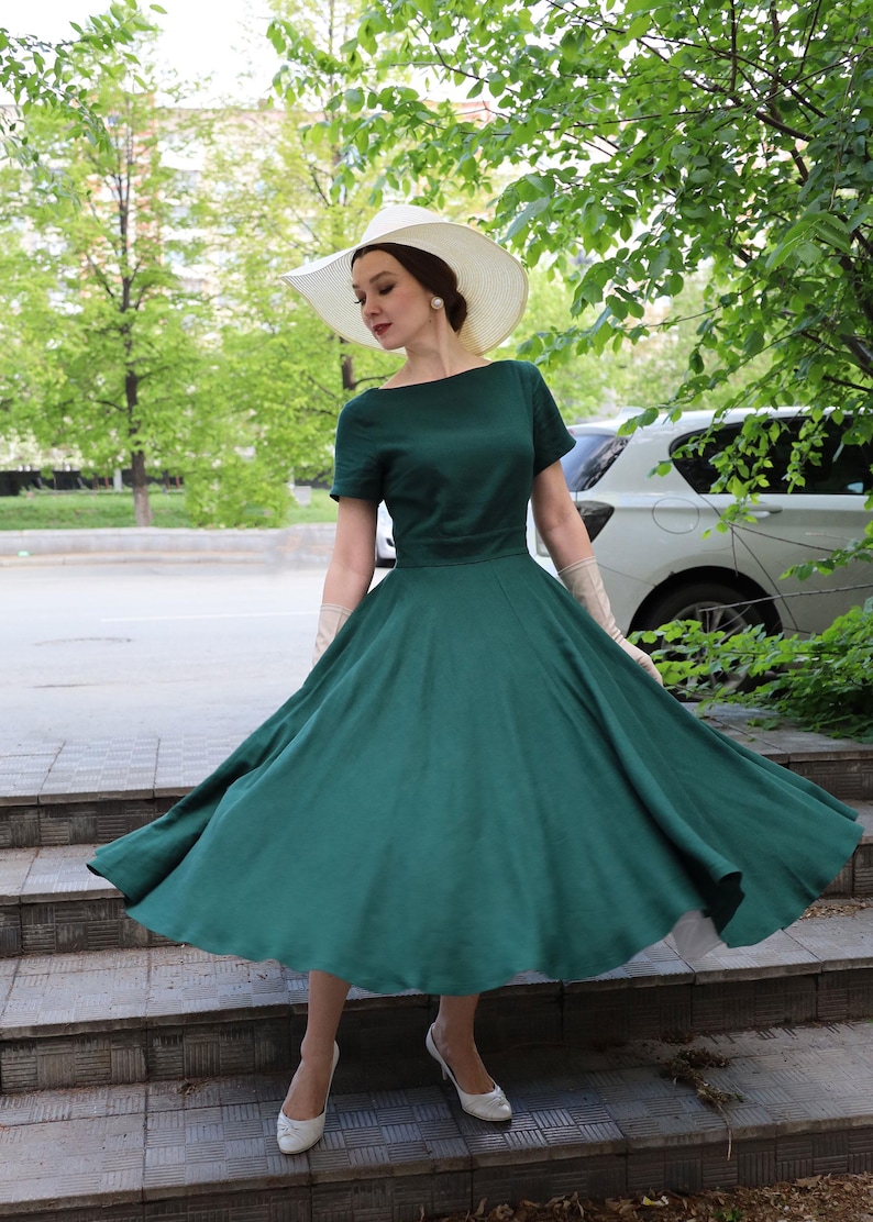 Puede incluir: Un vestido verde oscuro con escote barco y mangas cortas, que presenta un corpi&ntilde;o ajustado y una falda amplia y acampanada. El conjunto incluye un sombrero de sol blanco de ala ancha, guantes beige largos y zapatos de sal&oacute;n blancos cerrados.
