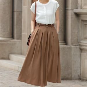 May include: A woman wearing a white short-sleeved blouse and a long brown pleated skirt. The outfit includes a brown belt, a brown shoulder bag, and black sandals. The background features a stone building.