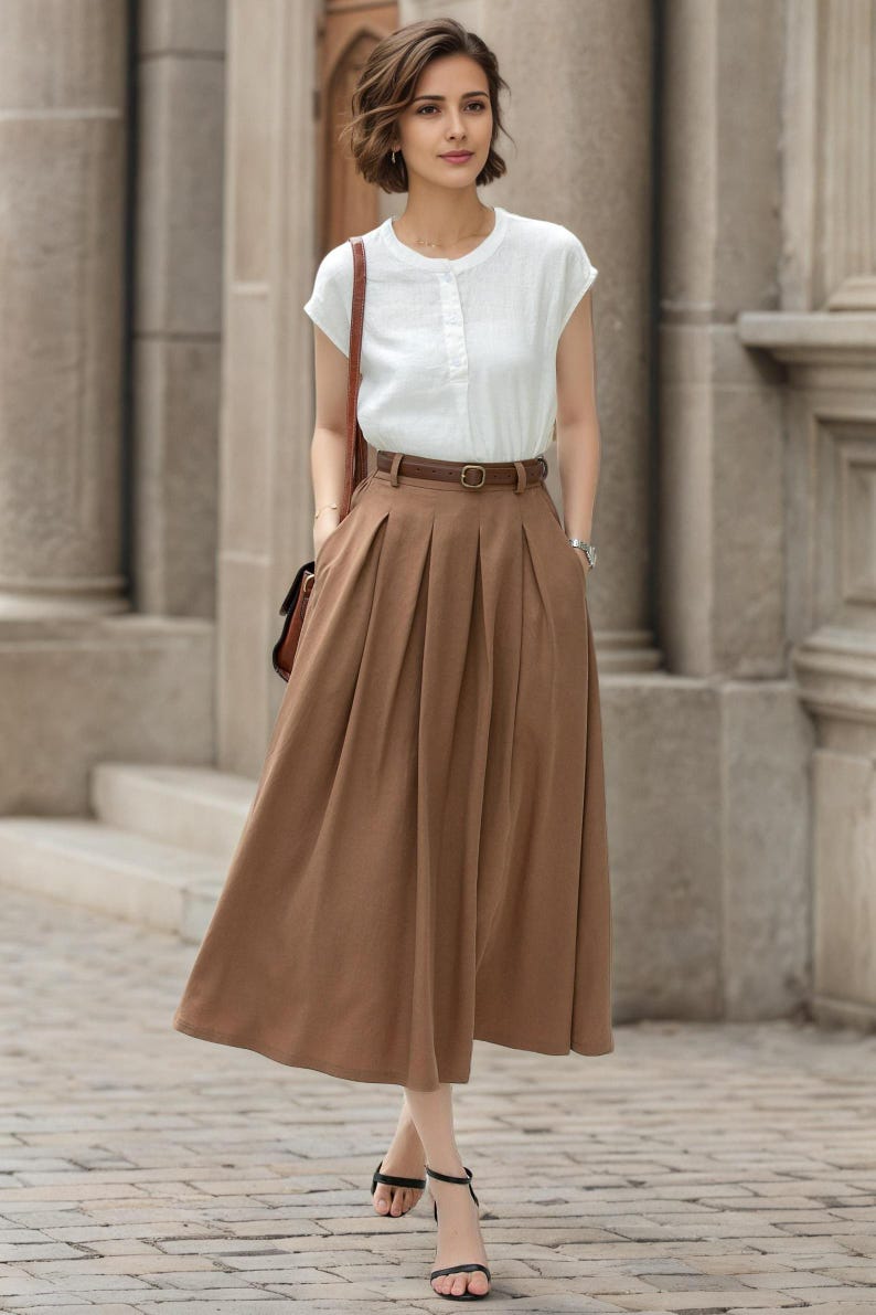 May include: A woman wearing a white short-sleeved blouse and a long brown pleated skirt. The outfit includes a brown belt, a brown shoulder bag, and black sandals. The background features a stone building.