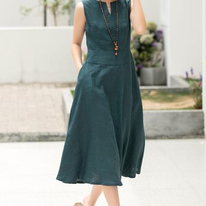 Peut inclure: Une robe en lin sans manches de couleur sarcelle avec un d&eacute;collet&eacute; en V et un collier de perles. La robe a une jupe ample et des poches sur les c&ocirc;t&eacute;s.