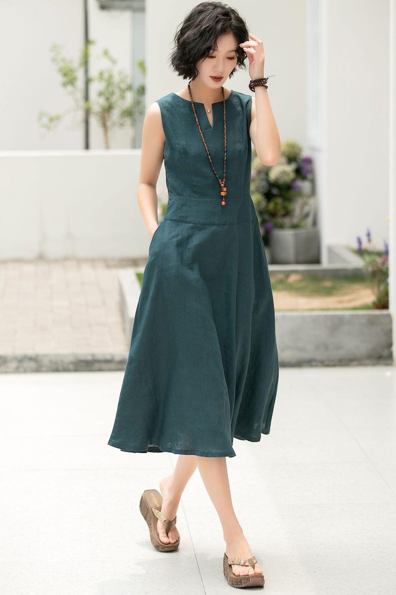 Peut inclure: Une robe en lin sans manches de couleur sarcelle avec un d&eacute;collet&eacute; en V et un collier de perles. La robe a une jupe ample et des poches sur les c&ocirc;t&eacute;s.