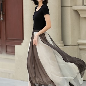 May include: A woman wearing a black short-sleeved top, a white hat, and a long, flowing black and white skirt. The skirt is sheer and has a layered design.