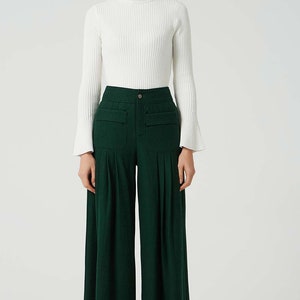 May include: A woman is wearing a white turtleneck sweater and dark green wide-leg trousers with pockets. The trousers have pleats and a button fastening.