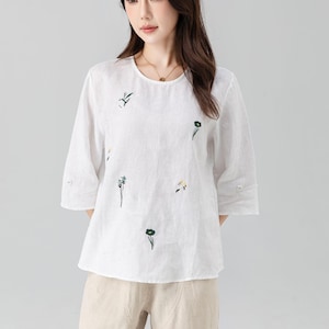 May include: White linen top with three-quarter sleeves and a round neckline. The blouse features delicate green floral embroidery. The model is wearing beige pants. This top is suitable for casual wear.