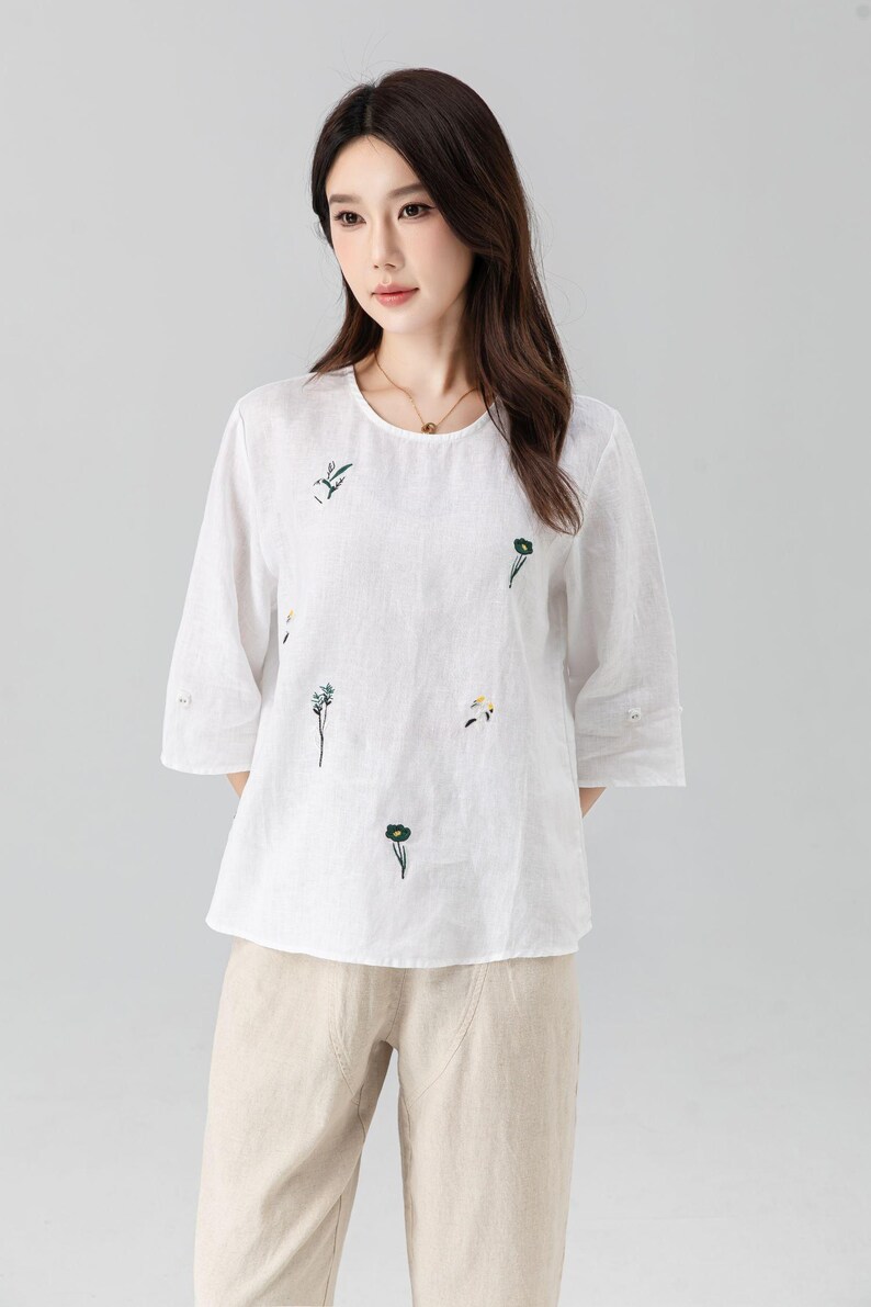 May include: White linen top with three-quarter sleeves and a round neckline. The blouse features delicate green floral embroidery. The model is wearing beige pants. This top is suitable for casual wear.