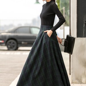May include: A woman wearing a black turtleneck sweater and a black and green plaid skirt. She is wearing a black hat with a safety pin design and black heeled boots. She is carrying a black handbag.
