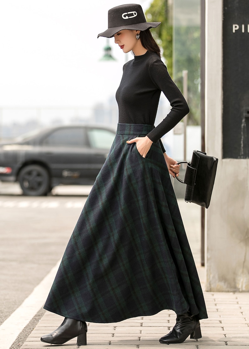 May include: A woman wearing a black turtleneck sweater and a black and green plaid skirt. She is wearing a black hat with a safety pin design and black heeled boots. She is carrying a black handbag.