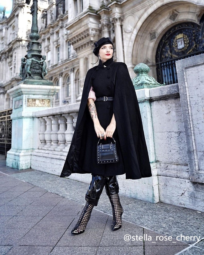 May include: A woman wearing a black cape, black boots, and a black beret with a small black purse. She is standing in front of a stone building with an archway.