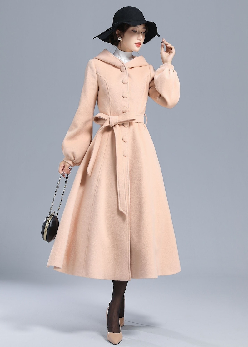 May include: A long, light pink coat with a hood, a belt, and a button closure. The coat has a flared skirt and long sleeves with puffed cuffs.
