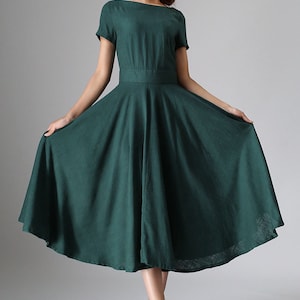 Puede incluir: Un vestido midi de color verde oscuro con escote barco, mangas cortas y una cintura entallada que se abre en una falda amplia y acampanada. La prenda est&aacute; confeccionada en una tela de color s&oacute;lido con textura visible y un dobladillo bajo la rodilla.