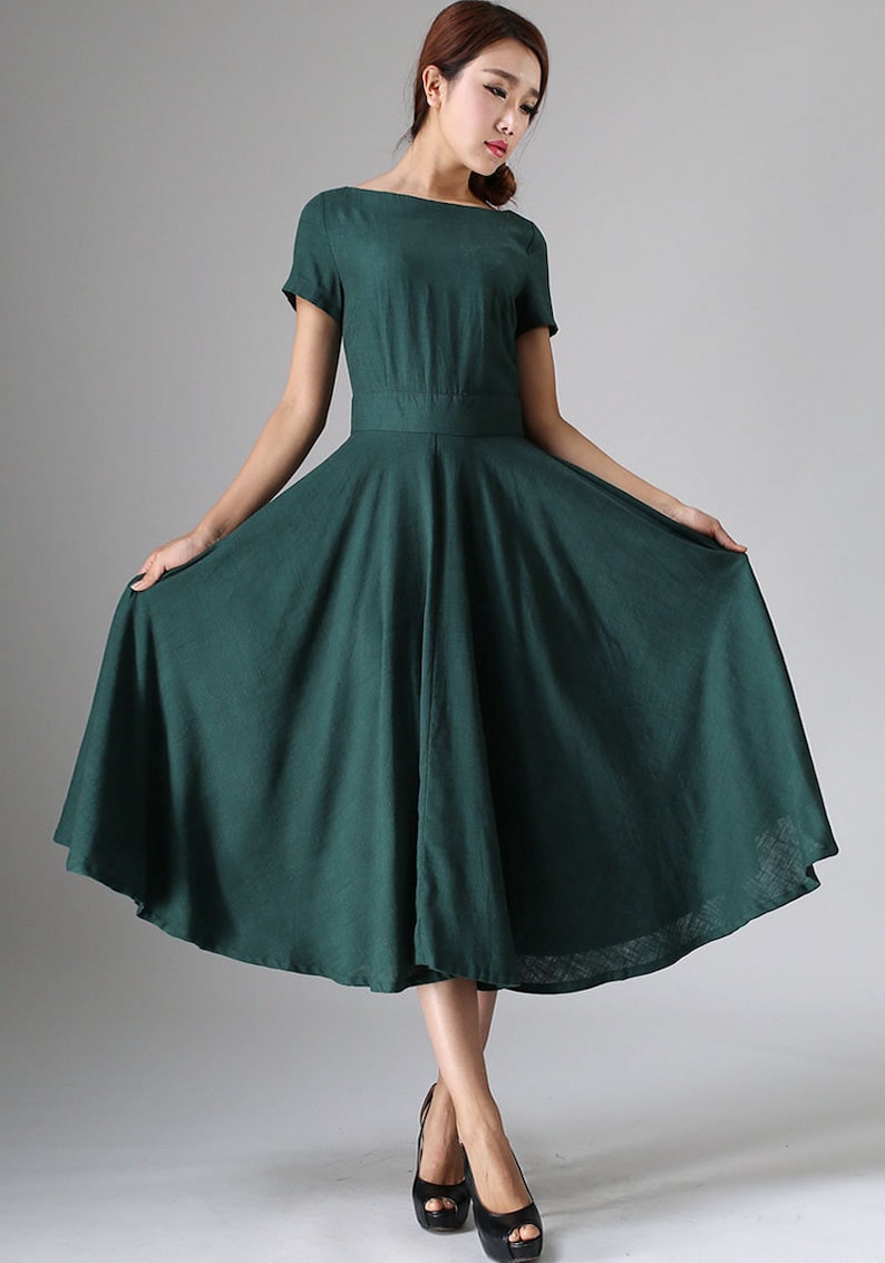 Puede incluir: Un vestido midi de color verde oscuro con escote barco, mangas cortas y una cintura entallada que se abre en una falda amplia y acampanada. La prenda est&aacute; confeccionada en una tela de color s&oacute;lido con textura visible y un dobladillo bajo la rodilla.