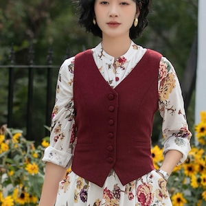 Puede incluir: Un chaleco burdeos con cuello en V y cierre de botones sobre una blusa y falda con estampado floral. La blusa es blanca con un colorido estampado floral. La falda tiene un diseño floral similar. El atuendo es adecuado para un día de primavera o verano.
