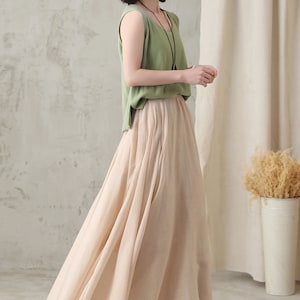 May include: A woman wearing a light green sleeveless top and a long, flowing beige skirt. The outfit is paired with beige slip-on shoes. The background is a neutral color, with a white curtain and a decorative vase.