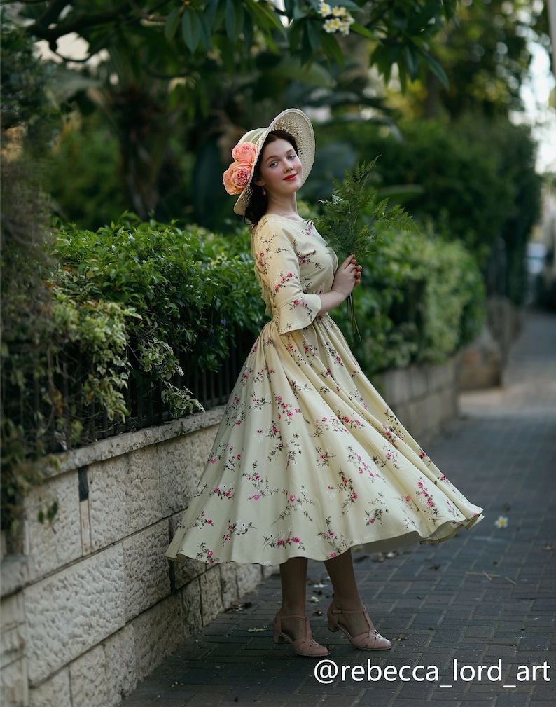 Peut inclure: Une robe vintage cr&egrave;me &agrave; motifs floraux roses, port&eacute;e avec un chapeau assorti orn&eacute; de roses roses. La robe a une jupe &eacute;vas&eacute;e et des manches trois-quarts. L'image est prise en ext&eacute;rieur.