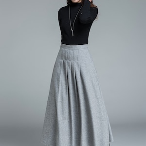 May include: A woman wearing a black turtleneck sweater and a long, gray pleated skirt. The skirt falls to the ankles. She is also wearing a silver necklace and black boots. The background is a solid gray.