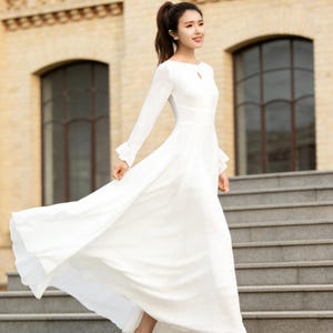 Puede incluir: Un vestido largo blanco con mangas largas y escote de ojo de cerradura. El vestido est&aacute; hecho de una tela ligera y se usa con tacones altos transparentes. La modelo camina por una escalera exterior.