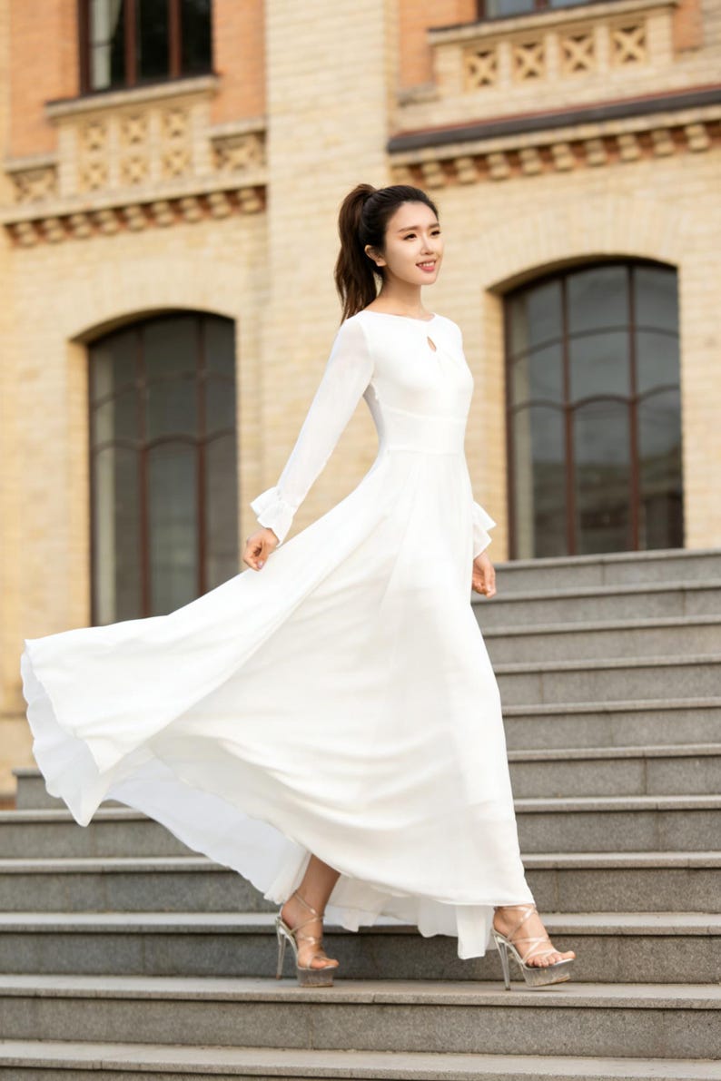 Puede incluir: Un vestido largo blanco con mangas largas y escote de ojo de cerradura. El vestido est&aacute; hecho de una tela ligera y se usa con tacones altos transparentes. La modelo camina por una escalera exterior.