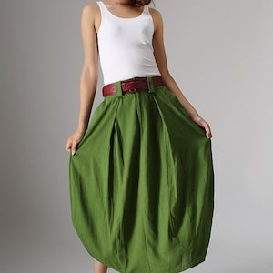 May include: A woman's outfit featuring a white tank top and a green skirt. The skirt is a vibrant green color and has a unique, rounded shape. A brown belt cinches the waist. The outfit is paired with sandals.