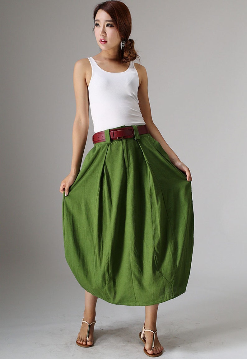 May include: A woman's outfit featuring a white tank top and a green skirt. The skirt is a vibrant green color and has a unique, rounded shape. A brown belt cinches the waist. The outfit is paired with sandals.