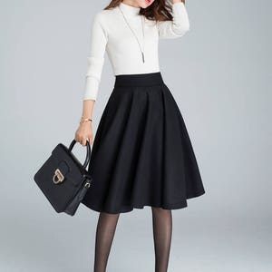 Peut inclure: Une femme portant un pull blanc &agrave; col roul&eacute; et une jupe noire taille haute. Elle porte un sac &agrave; main noir et des collants noirs et des talons.