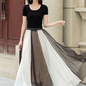 May include: A woman wearing a black short-sleeved top and a white and black sheer maxi skirt. She is wearing a straw hat and has her hair pulled back. She is standing in front of a white wall.