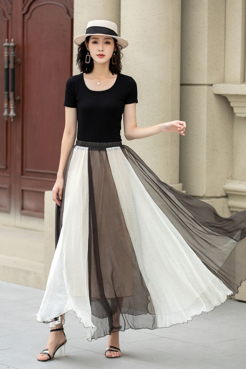 May include: A woman wearing a black short-sleeved top and a white and black sheer maxi skirt. She is wearing a straw hat and has her hair pulled back. She is standing in front of a white wall.