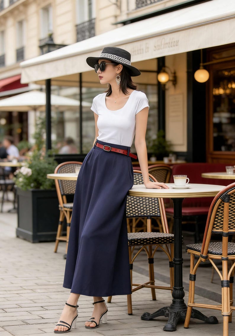 May include: A woman wearing a white short-sleeve top, a navy blue maxi skirt, and a red belt. She is wearing a black hat and sunglasses. She is standing near a cafe table with a cup of coffee.