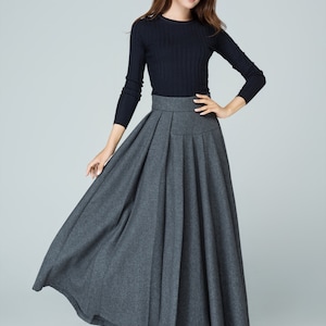 May include: A woman wearing a black long-sleeved sweater and a gray pleated maxi skirt. She is standing on a white background.