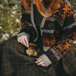 Pu&ograve; includere: Una donna indossa una gonna a quadri marroni e rossi con un cardigan a motivi marroni, rossi e gialli. Tiene in mano alcune foglie marroni.