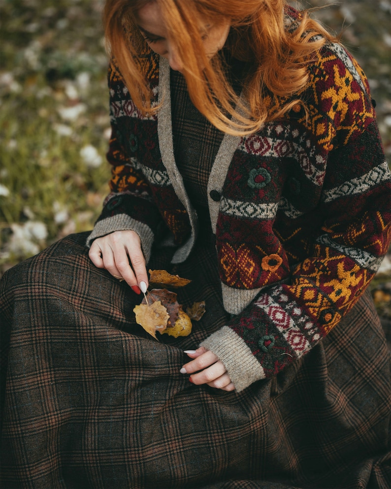 Pu&ograve; includere: Una donna indossa una gonna a quadri marroni e rossi con un cardigan a motivi marroni, rossi e gialli. Tiene in mano alcune foglie marroni.