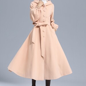 May include: A long, blush pink coat with a hood, a belt, and button closures. The coat has a flared skirt and long sleeves with puffed cuffs.