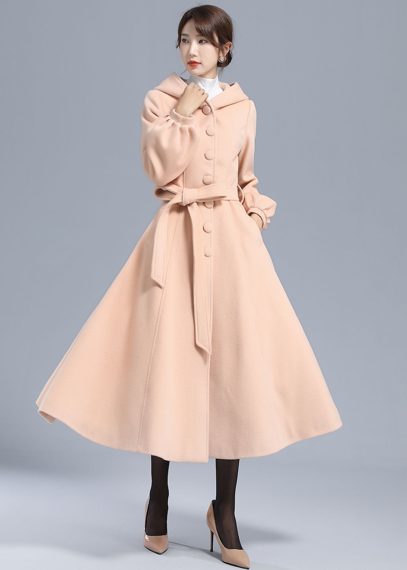 May include: A long, blush pink coat with a hood, a belt, and button closures. The coat has a flared skirt and long sleeves with puffed cuffs.