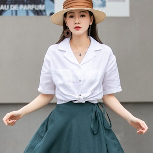 May include: A woman wearing a white linen shirt with a knotted front and a dark green wrap skirt. She is wearing a wide-brimmed straw hat with a brown band and brown geometric earrings.