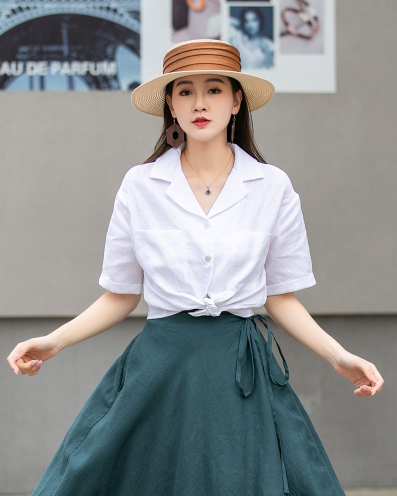 May include: A woman wearing a white linen shirt with a knotted front and a dark green wrap skirt. She is wearing a wide-brimmed straw hat with a brown band and brown geometric earrings.