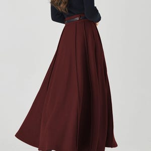 May include: A burgundy-colored, pleated midi skirt paired with a black long-sleeve top and a black belt. The outfit is completed with black high-heeled shoes. The skirt has a flowing, A-line silhouette.