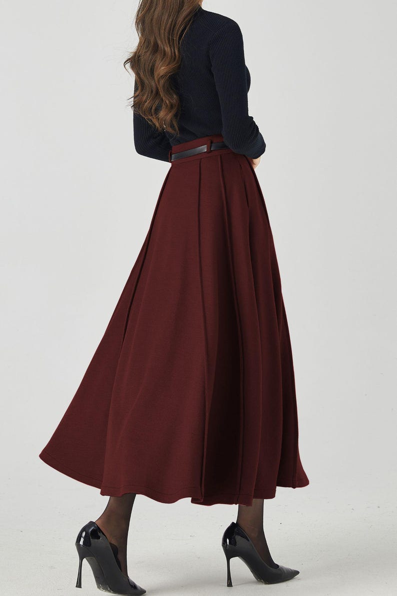 May include: A burgundy-colored, pleated midi skirt paired with a black long-sleeve top and a black belt. The outfit is completed with black high-heeled shoes. The skirt has a flowing, A-line silhouette.