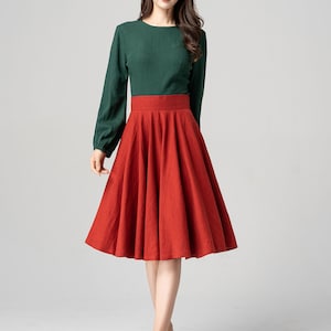 May include: A woman wearing a green long-sleeved shirt and a red A-line skirt. She is standing on a white background.