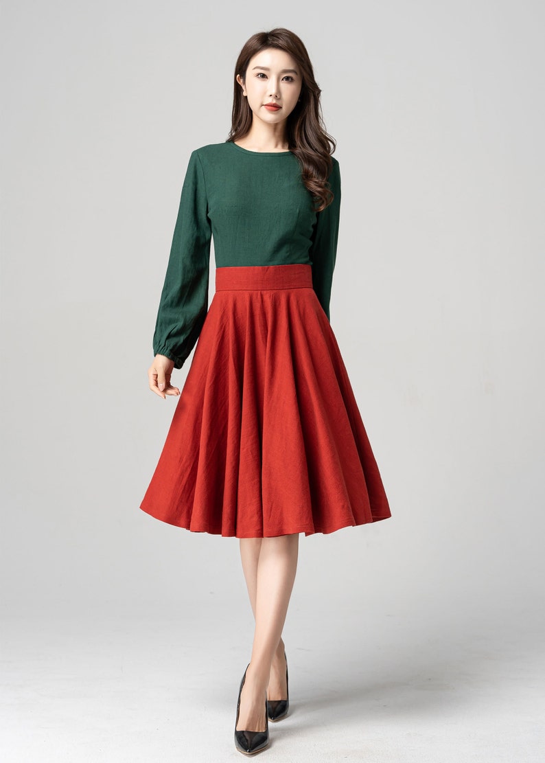 May include: A woman wearing a green long-sleeved shirt and a red A-line skirt. She is standing on a white background.