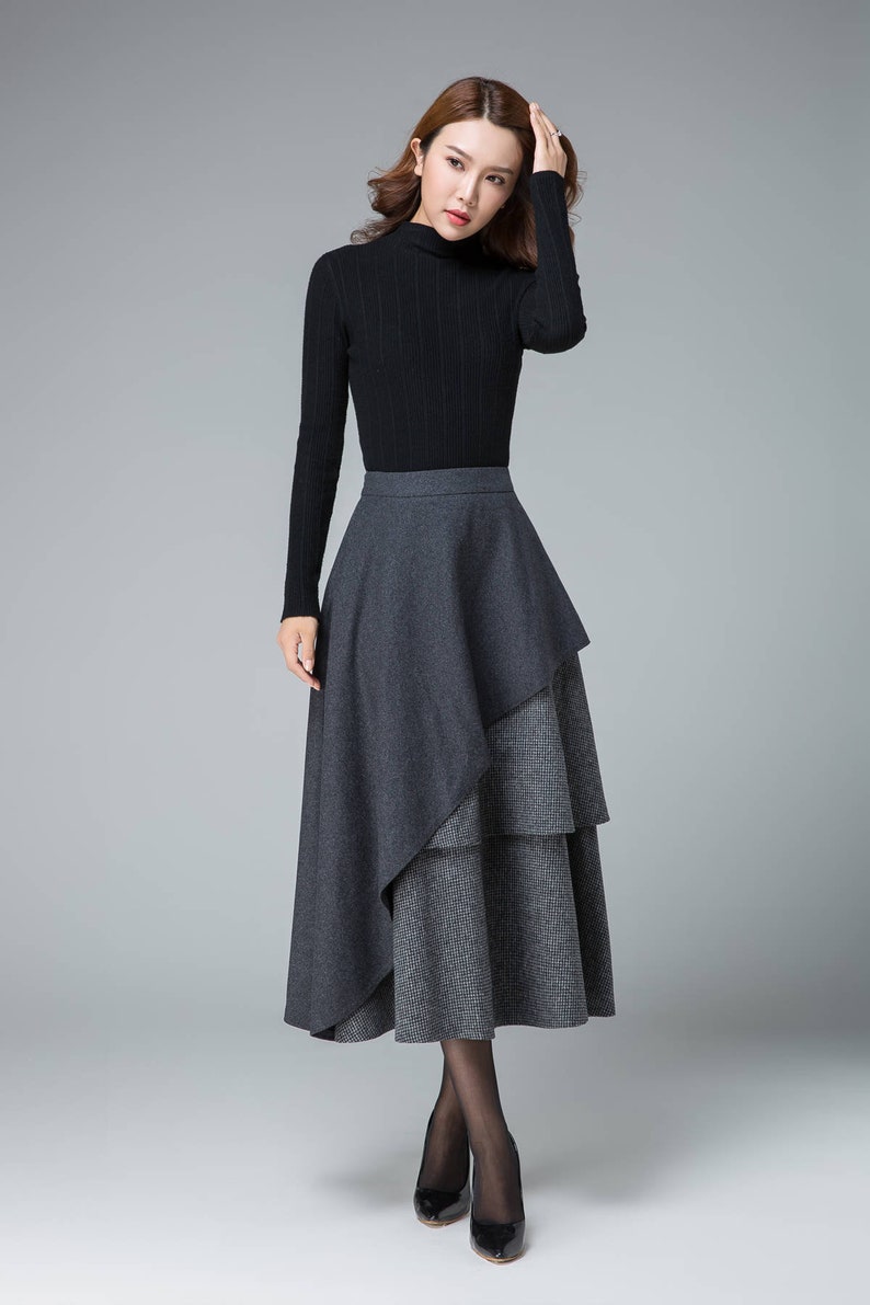 May include: A woman wearing a black turtleneck sweater and a gray tiered skirt with a black and white plaid pattern. The skirt is asymmetrical and has a longer layer in the front.